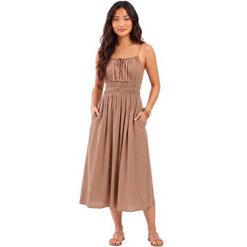 A woman modeling a sleeveless midi dress in a warm taupe-brown color, featuring a gathered smocked bodice, thin shoulder straps, and side pockets. The brand name 'AURA' is printed in black text on the left side of the image, set against a solid pale beige background.
