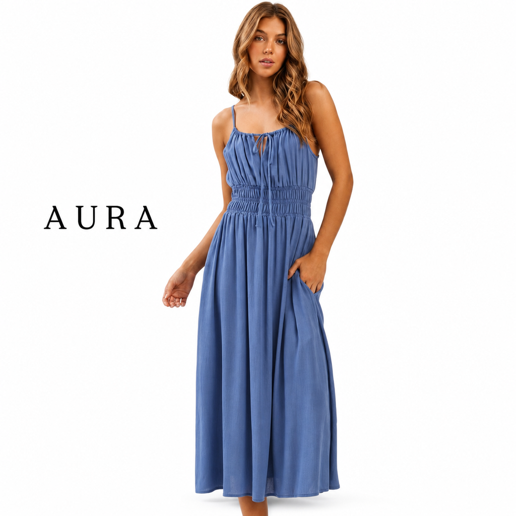 A woman modeling a sleeveless midi dress in dusty blue, featuring thin spaghetti straps, a smocked bodice, and side pockets. The brand name 'AURA' is printed in black serif font on the left side of the image, set against a plain white background.