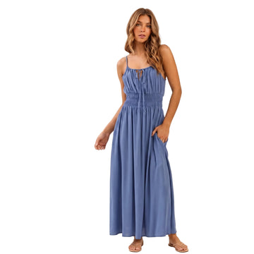A woman modeling a sleeveless, floor-length blue maxi dress with a smocked waist and front keyhole tie detail, shown against a plain light beige studio background. The brand name AURA appears in black text on the left side.